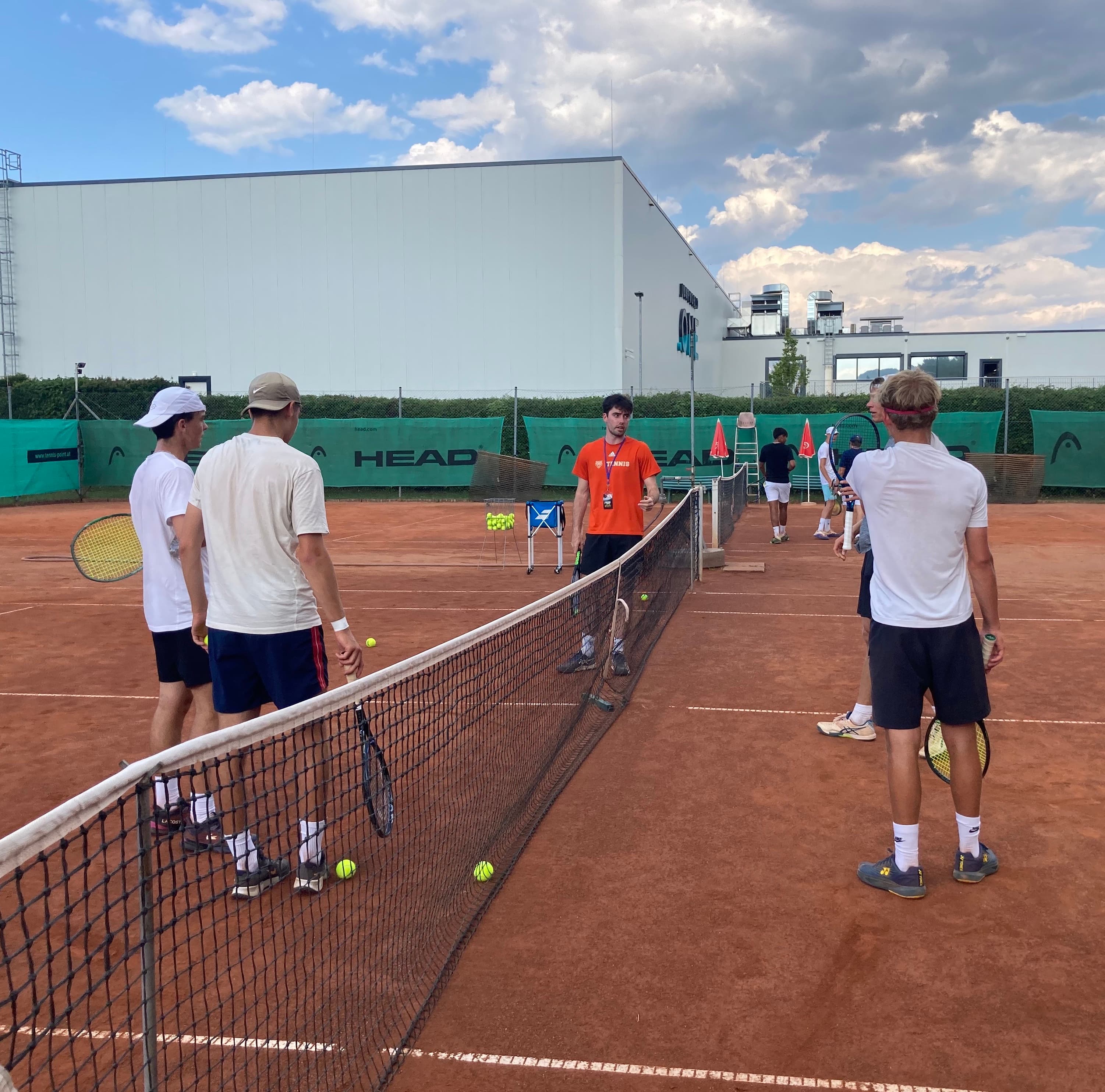 Eine Gruppe Tennisspieler steht während einer Trainingseinheit auf einem roten Sandplatz und hört einem US-College-Trainer im orangefarbenen Shirt zu.