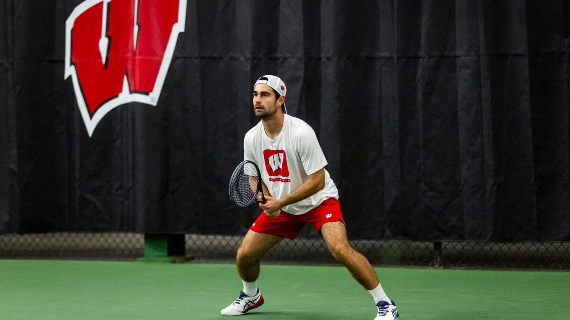 Gabriel Huber Playing College Tennis at the University of Wisconsin - Madison College tennis player Gabriel Huber from the University of Wisconsin - Madison focused and waiting for the ball to come to his side of the court during a match.